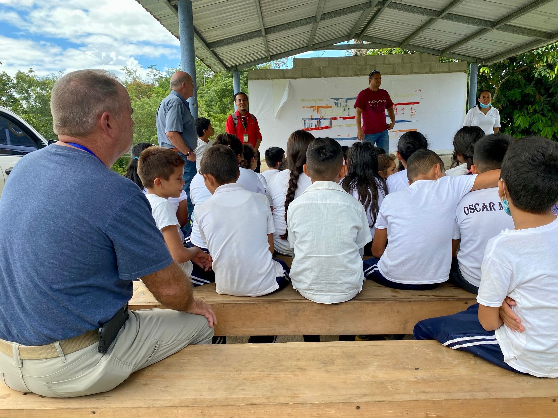 A group of children are sitting on a wooden bench under a canopy.