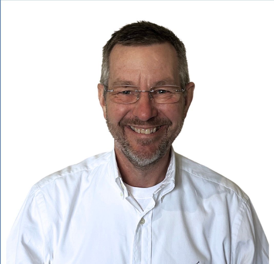 Man with glasses smiles, wearing white shirt, against a light background.