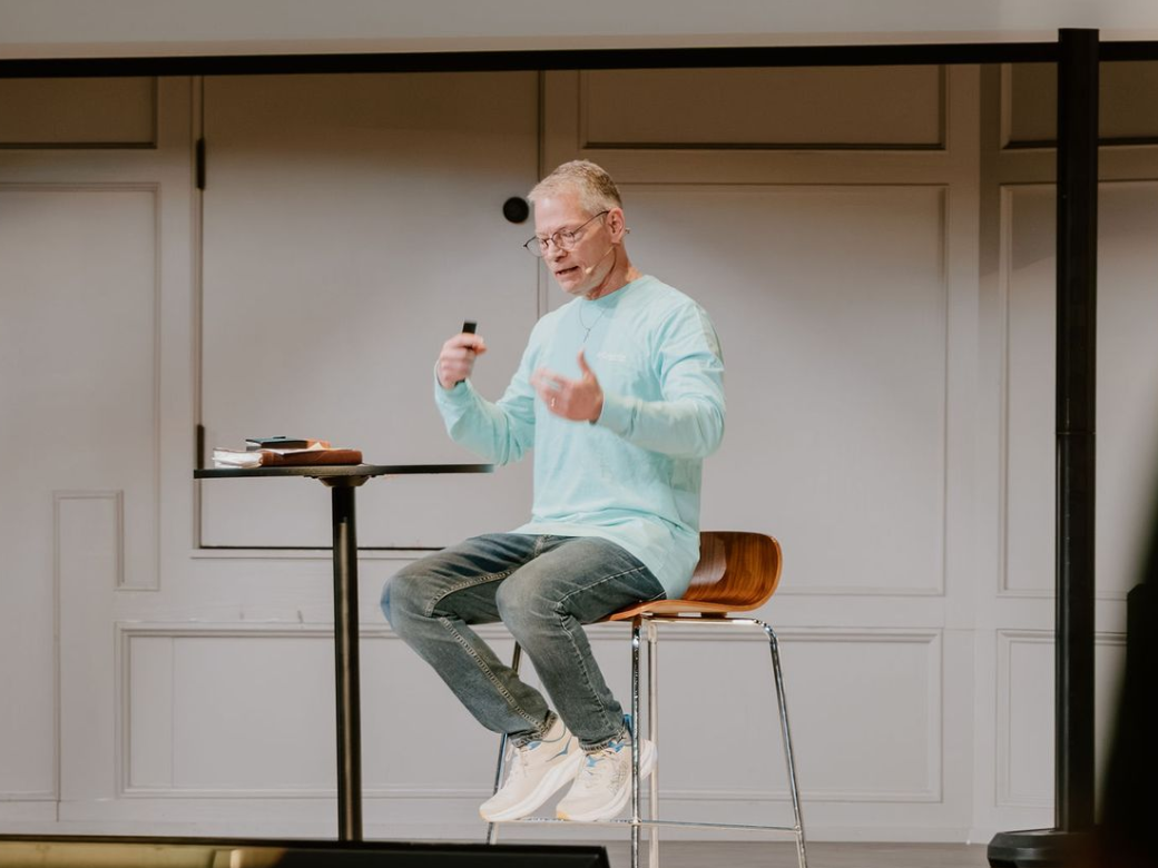 Man speaking on stage beside a small table, wearing a light blue shirt and jeans, holding a microphone.