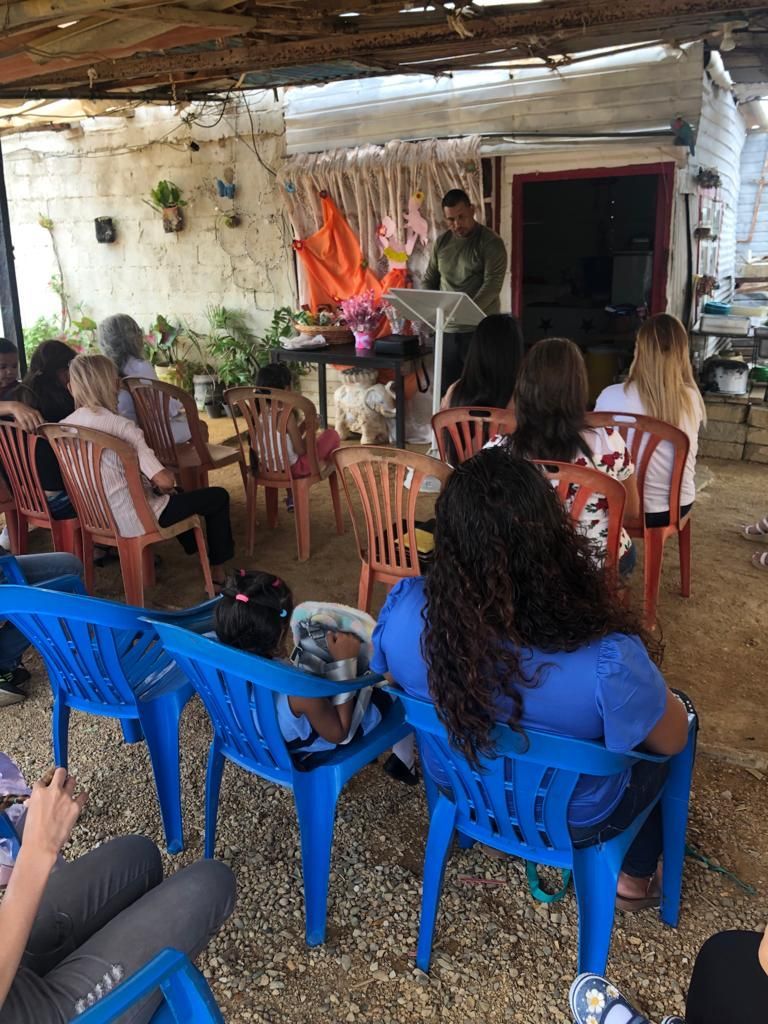 A group of people are sitting in blue chairs in front of a man at a podium.