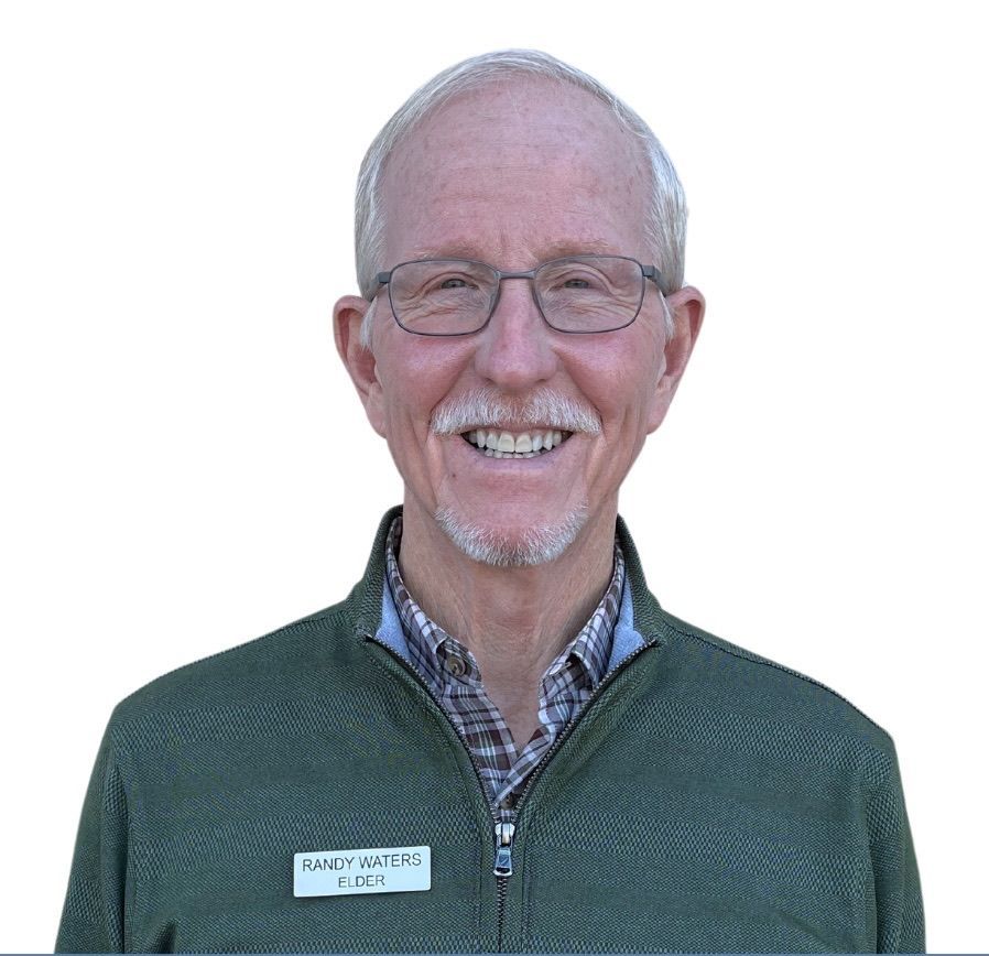 Smiling older man with glasses, name tag, green sweater, and blue-and-white collared shirt.