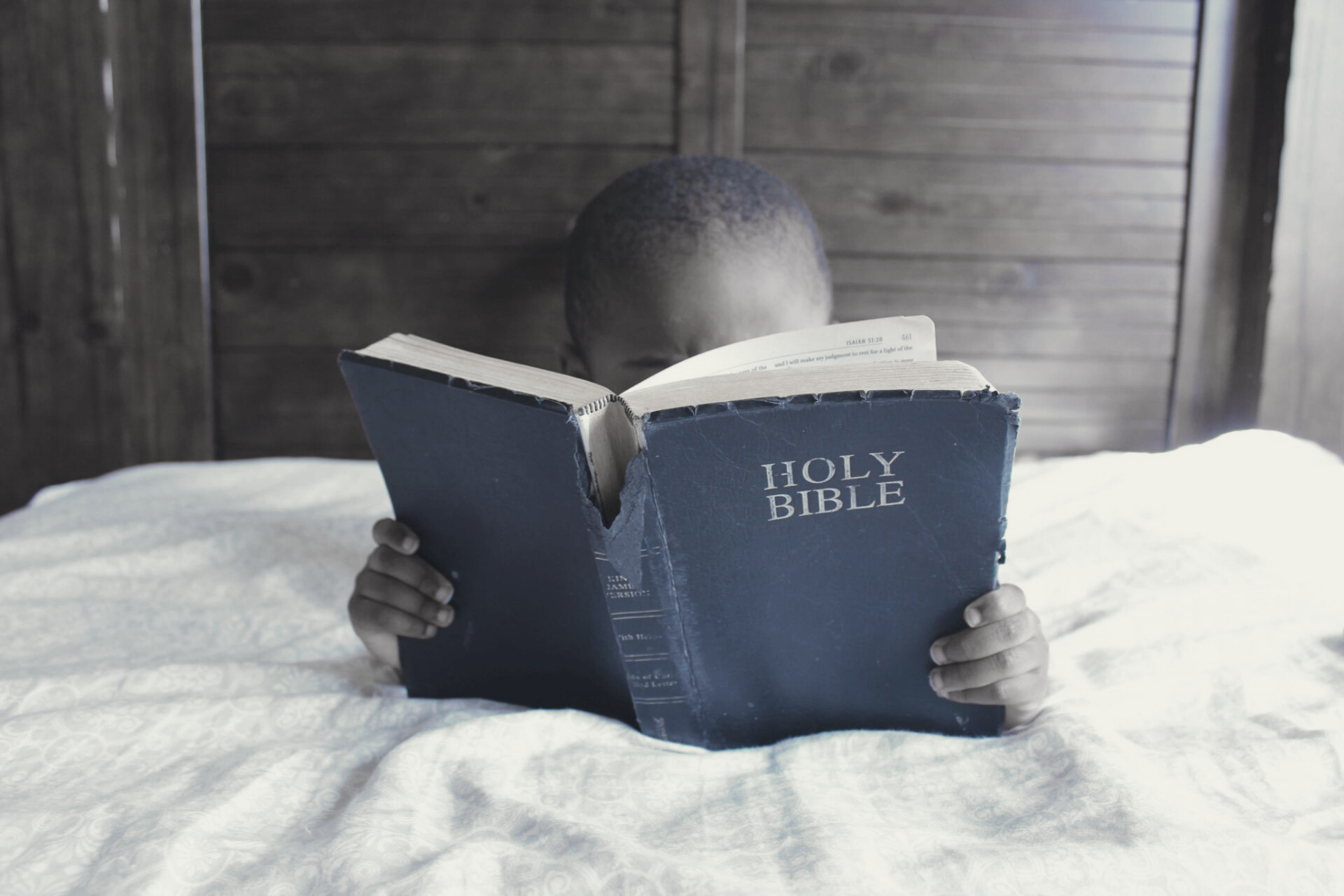 A child is laying on a bed reading a holy bible.