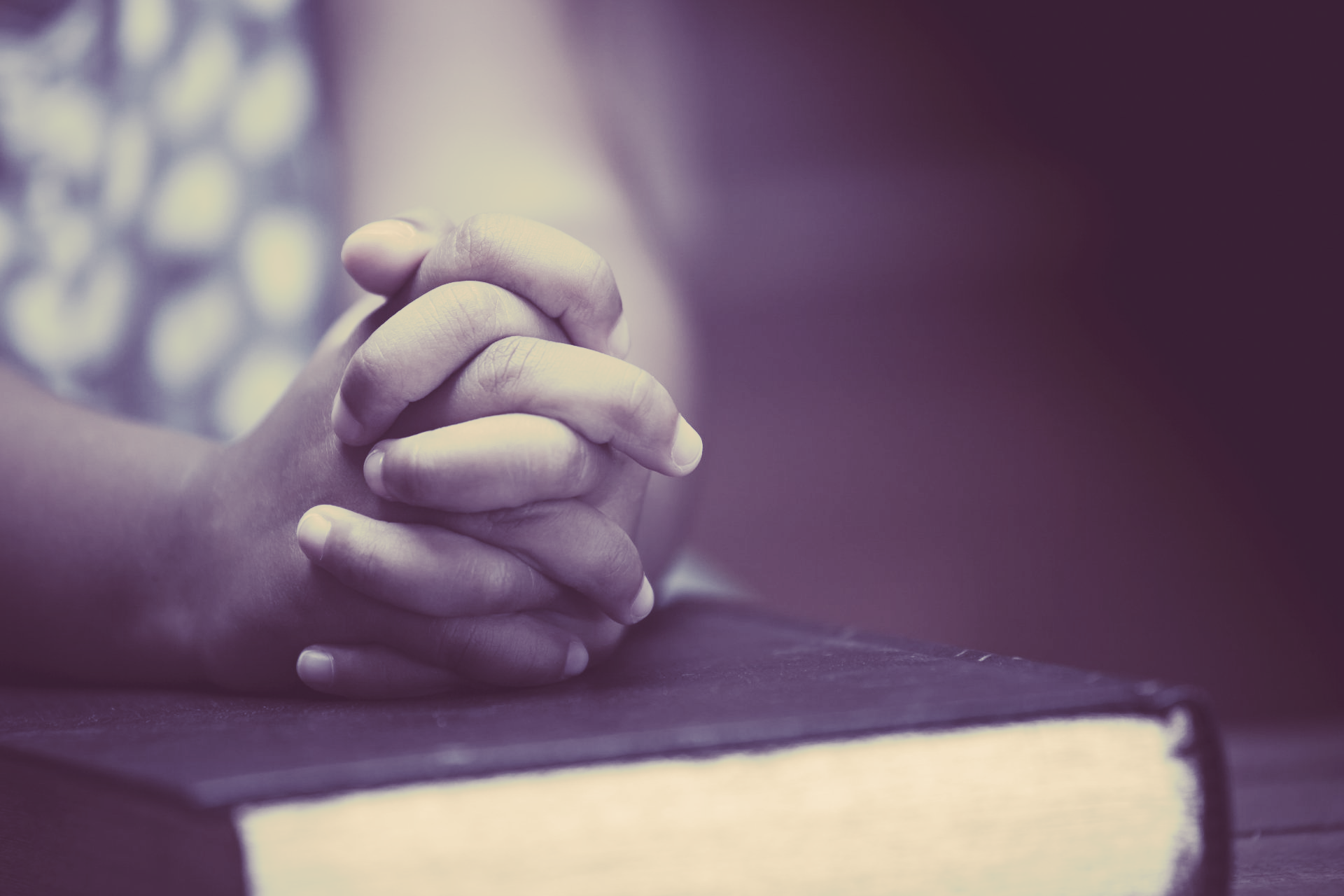 A person is praying with their hands folded over a bible.
