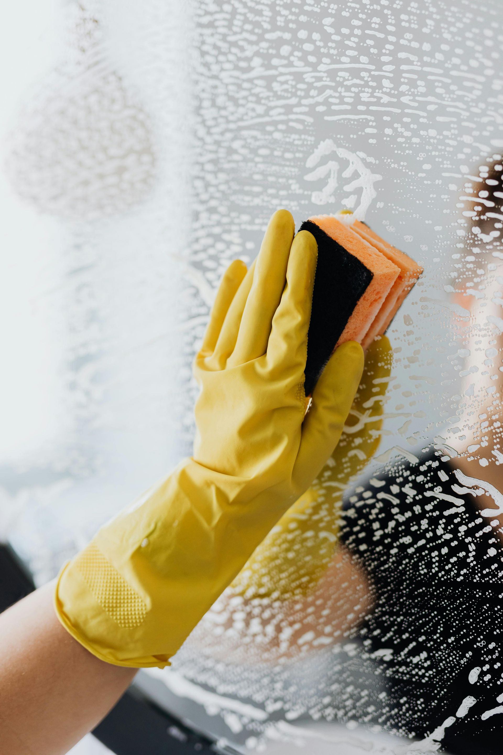 Yellow-gloved hand cleaning a soapy mirror with a sponge.