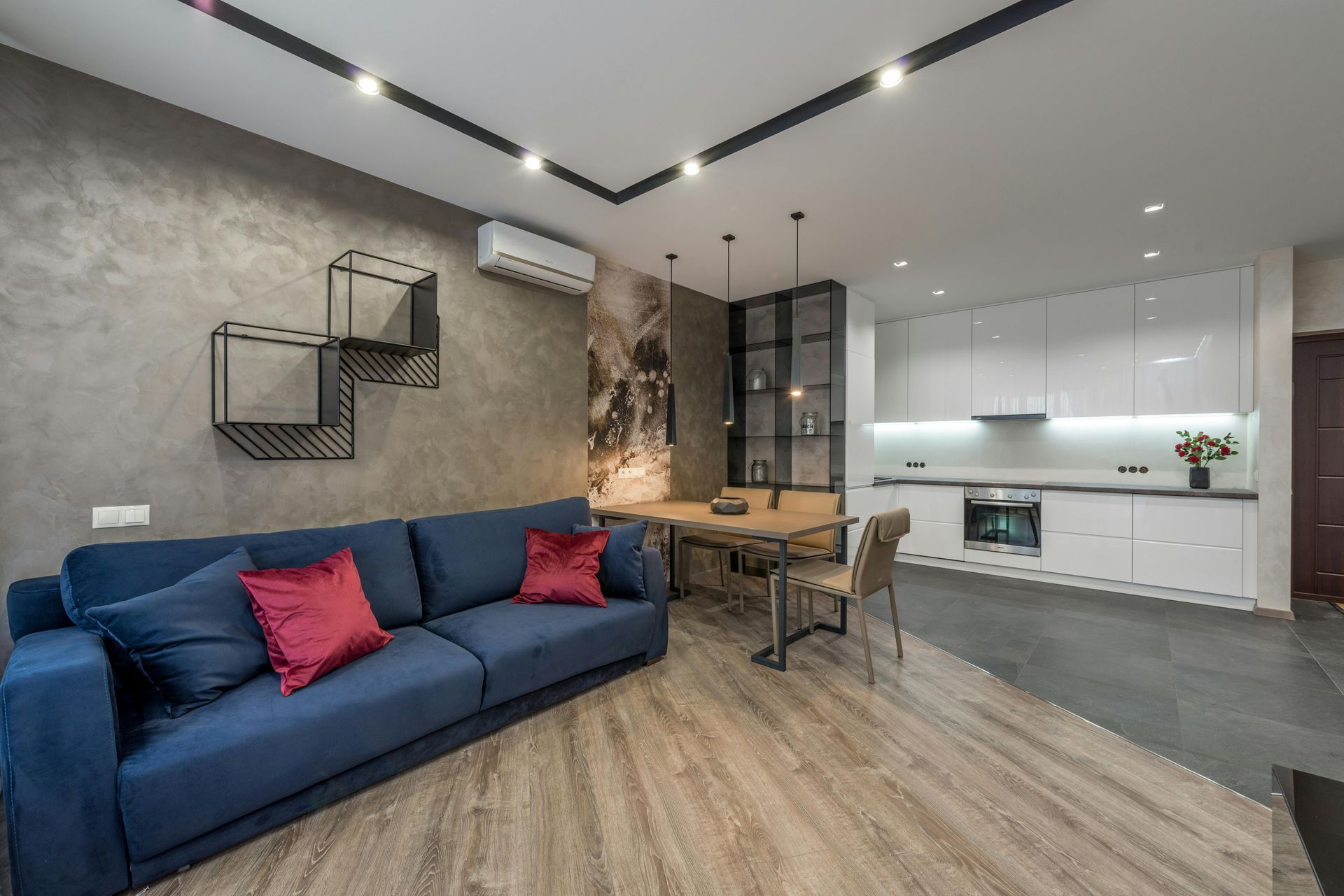 Modern open-plan living space with blue sofa, dining area, and white kitchen. Grey walls and light-colored wood flooring.