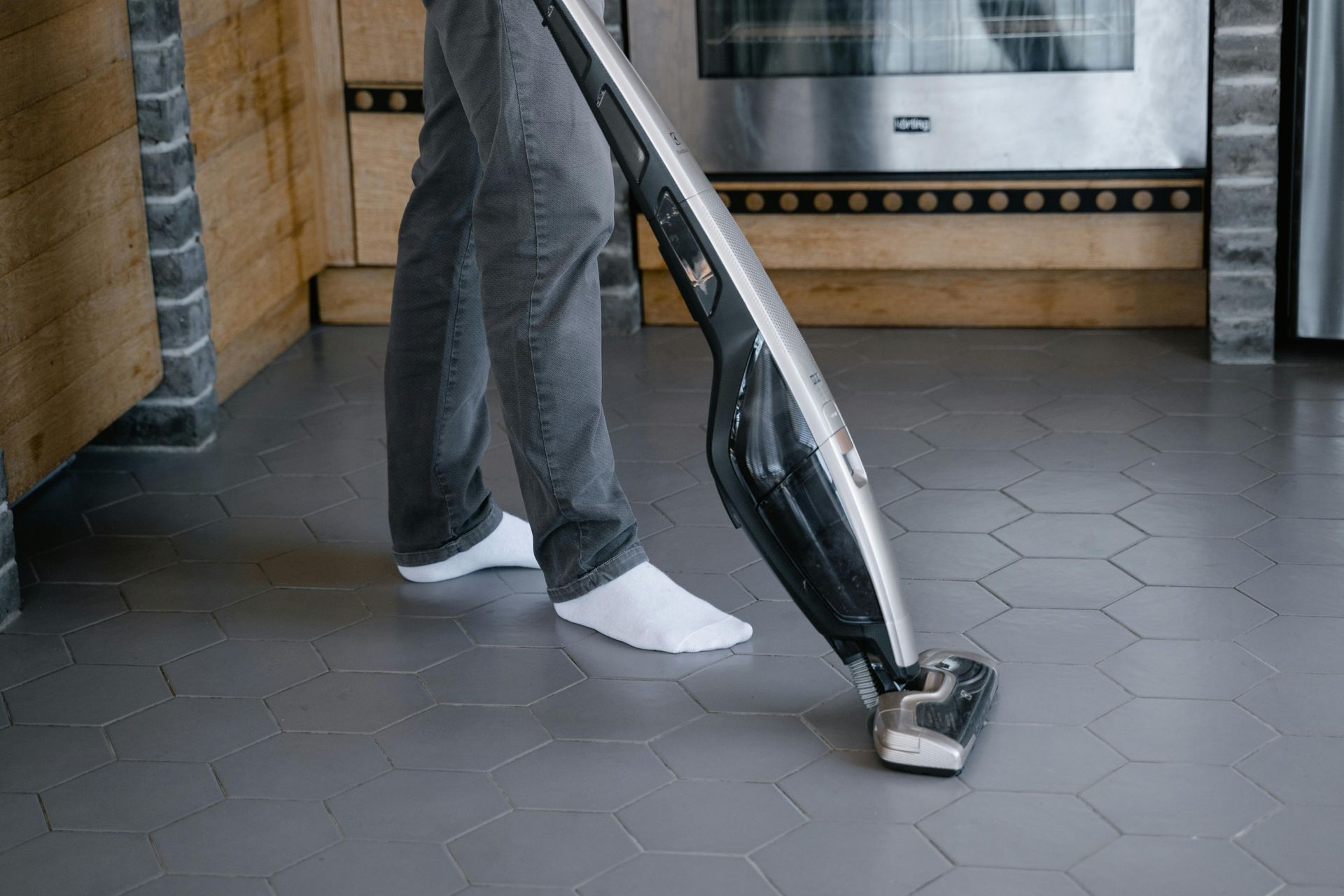 Person vacuums a gray hexagonal tiled floor with a black and silver stick vacuum.