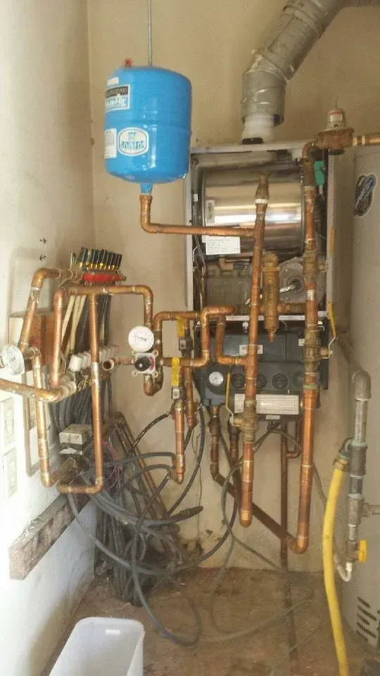 Wall-mounted gas boiler with copper pipes, a blue expansion tank, and a vent pipe in a small room.