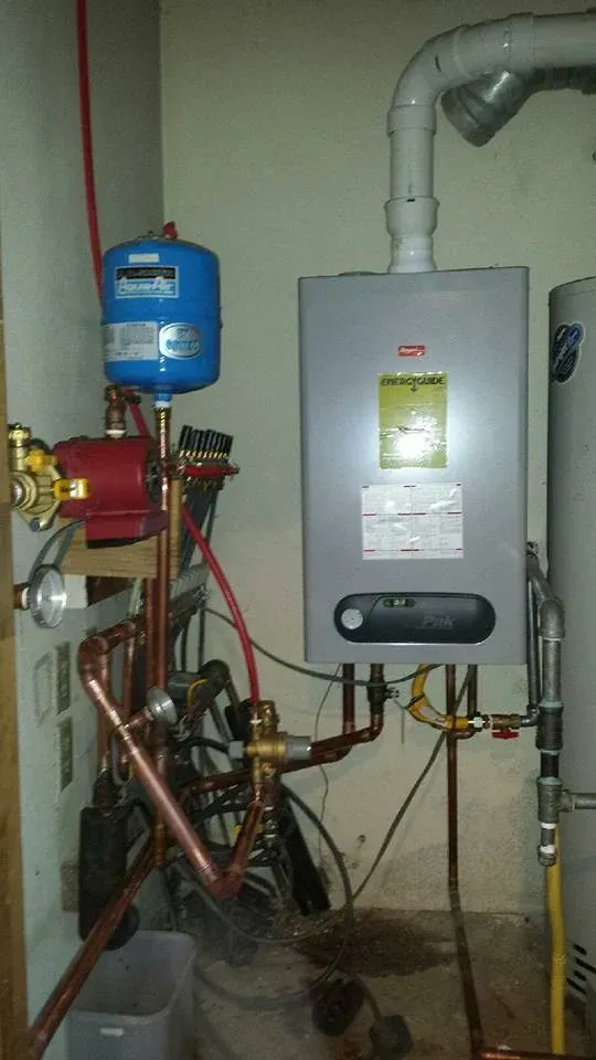 Water heater and related plumbing in a basement, featuring copper pipes, a red pump, and a blue expansion tank.