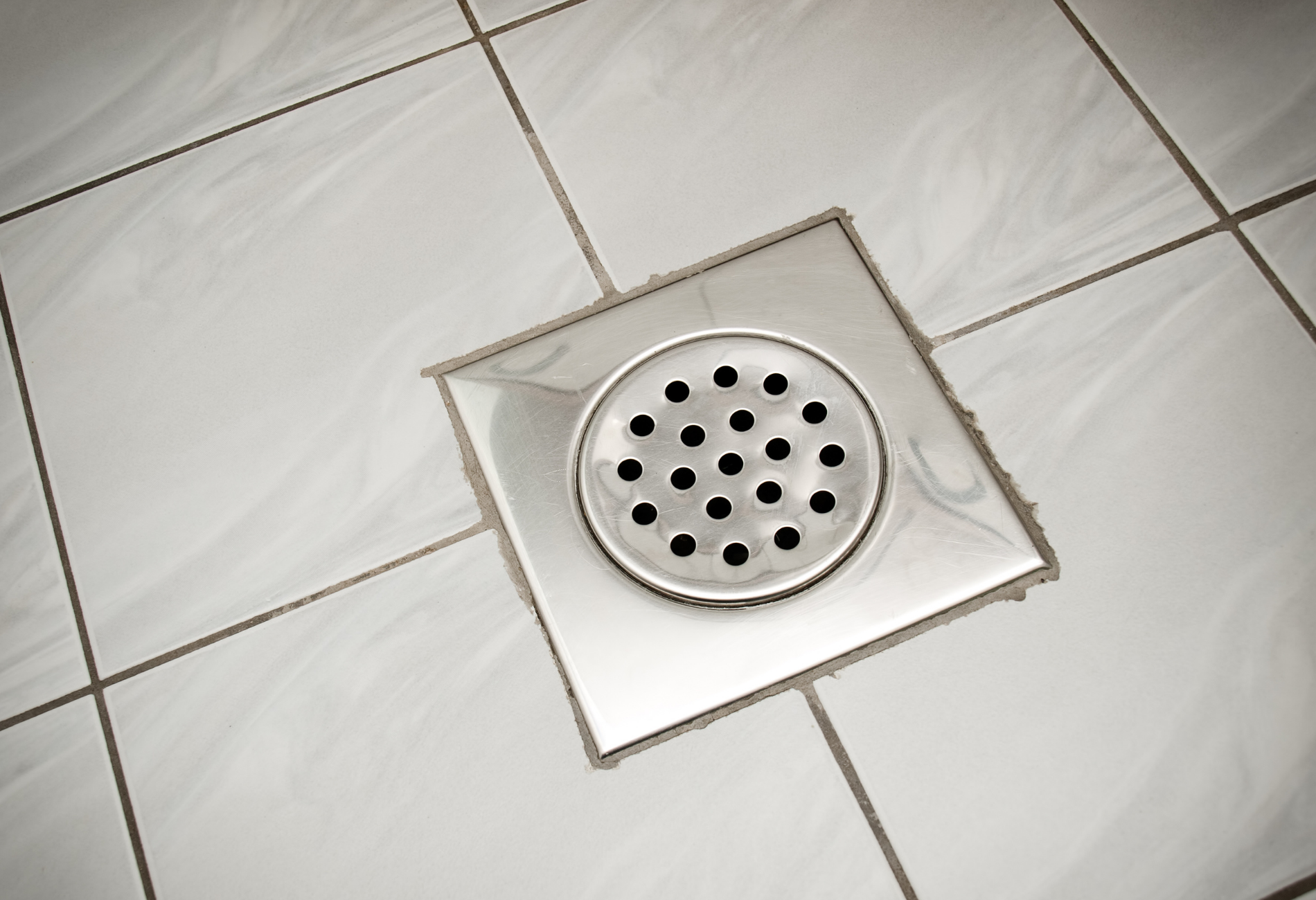 Square, stainless steel shower drain in white tiled floor.