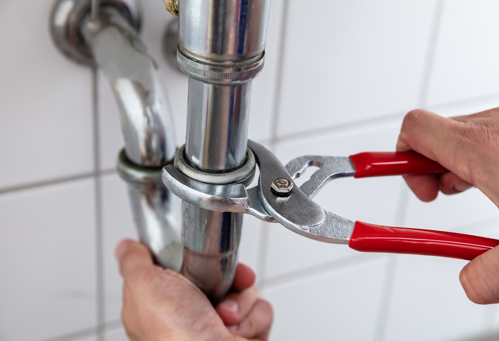Plumber using pliers to tighten a silver pipe under a white tiled wall. Red handled pliers.