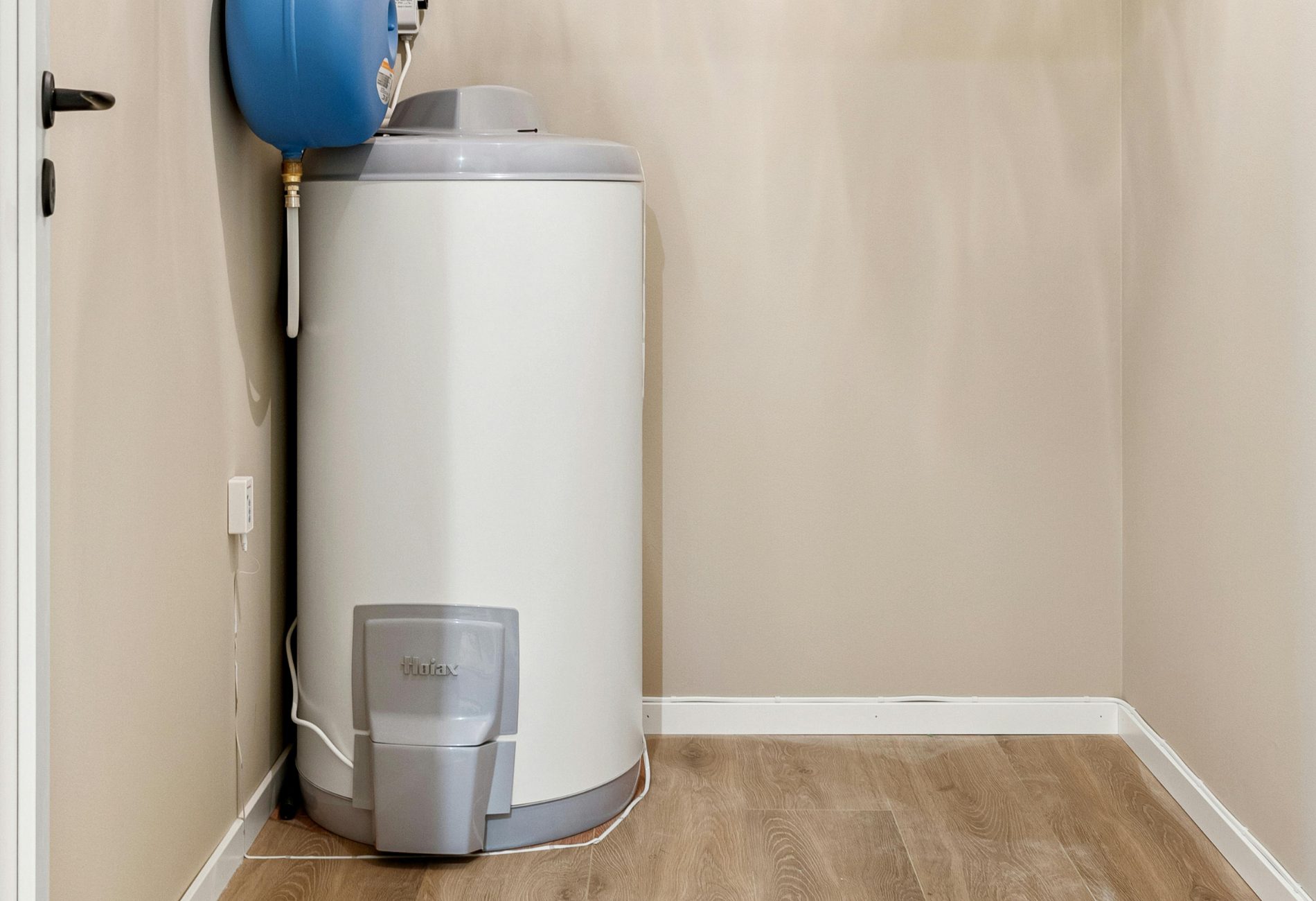 Water heater in a small room, with a blue expansion tank on top. Beige walls and wood-look floor.