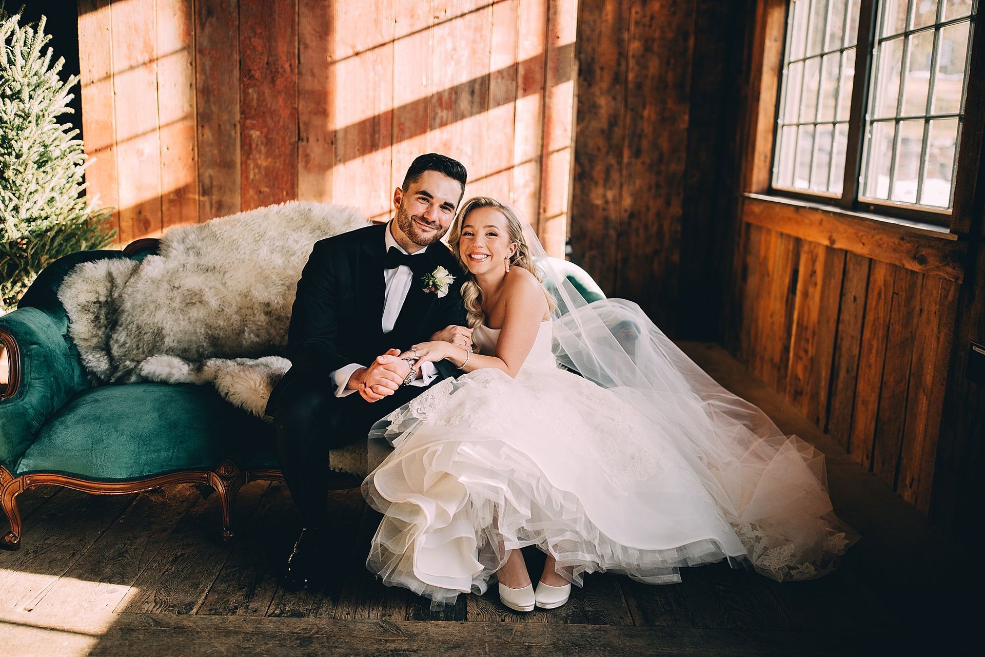 Wedding couple on a teal sofa, man in tuxedo, woman in a white dress, inside a wooden building.