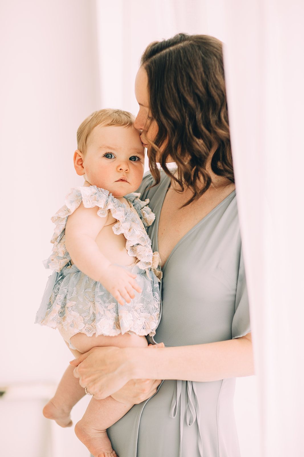 Woman in gray dress holding a baby wearing a lace outfit; woman kisses baby's forehead.