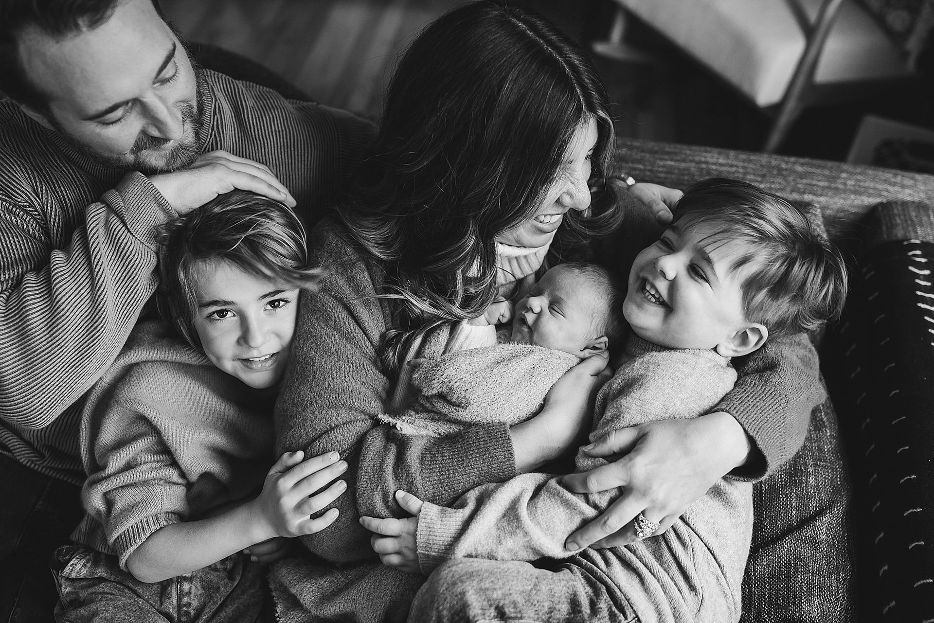 Family embraces: mother holds a baby and a laughing toddler, with another child and father nearby.