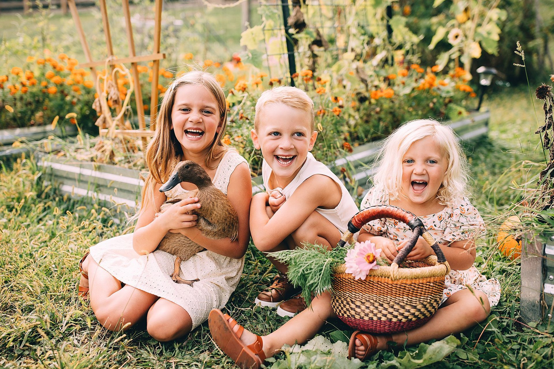 Three children laughing in a garden, one holding a chicken, another with a basket of flowers, surrounded by plants and blooms.