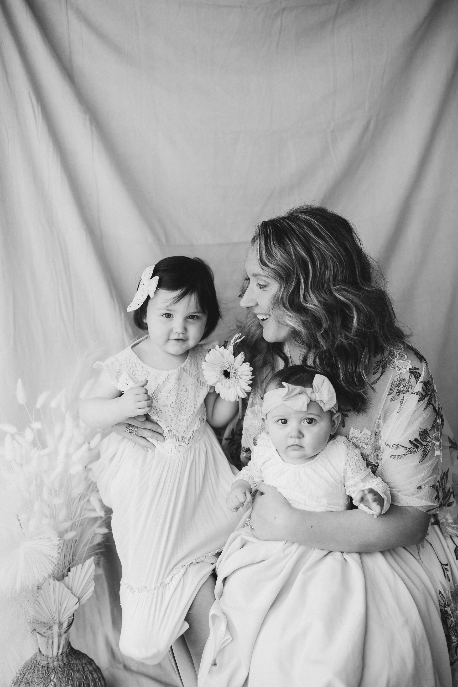 Woman sitting with two children, all wearing floral headbands and dresses, holding a flower.