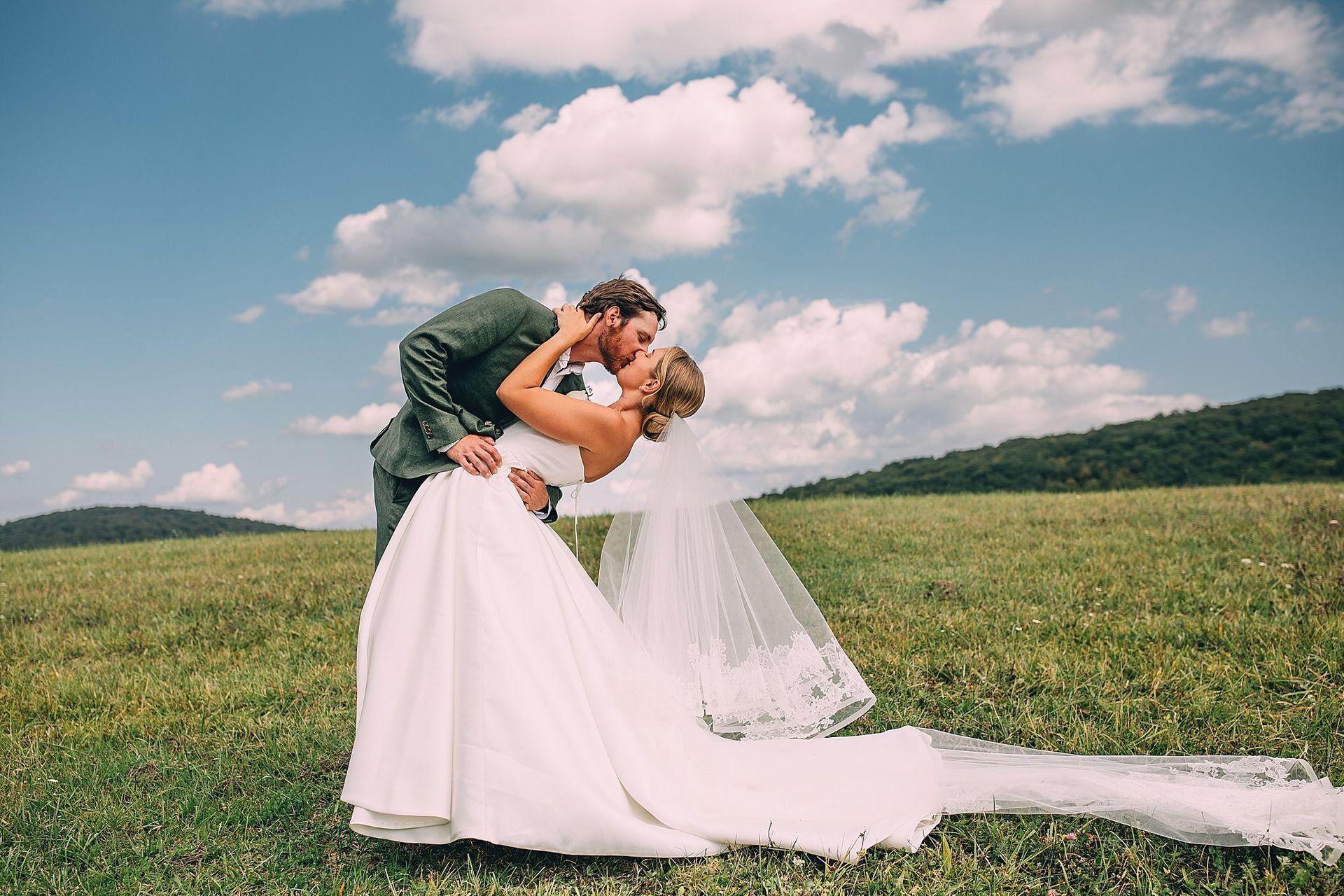 Couple kissing in a field, woman in a white wedding dress and veil, man in a green suit. Blue sky with clouds.