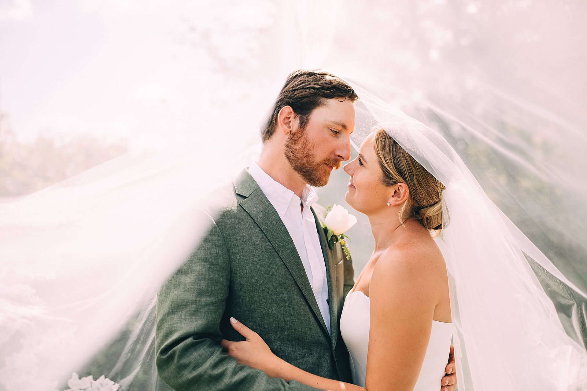 Bride and groom embrace, under sheer veil, outdoor setting. Groom in green suit, bride in white dress.