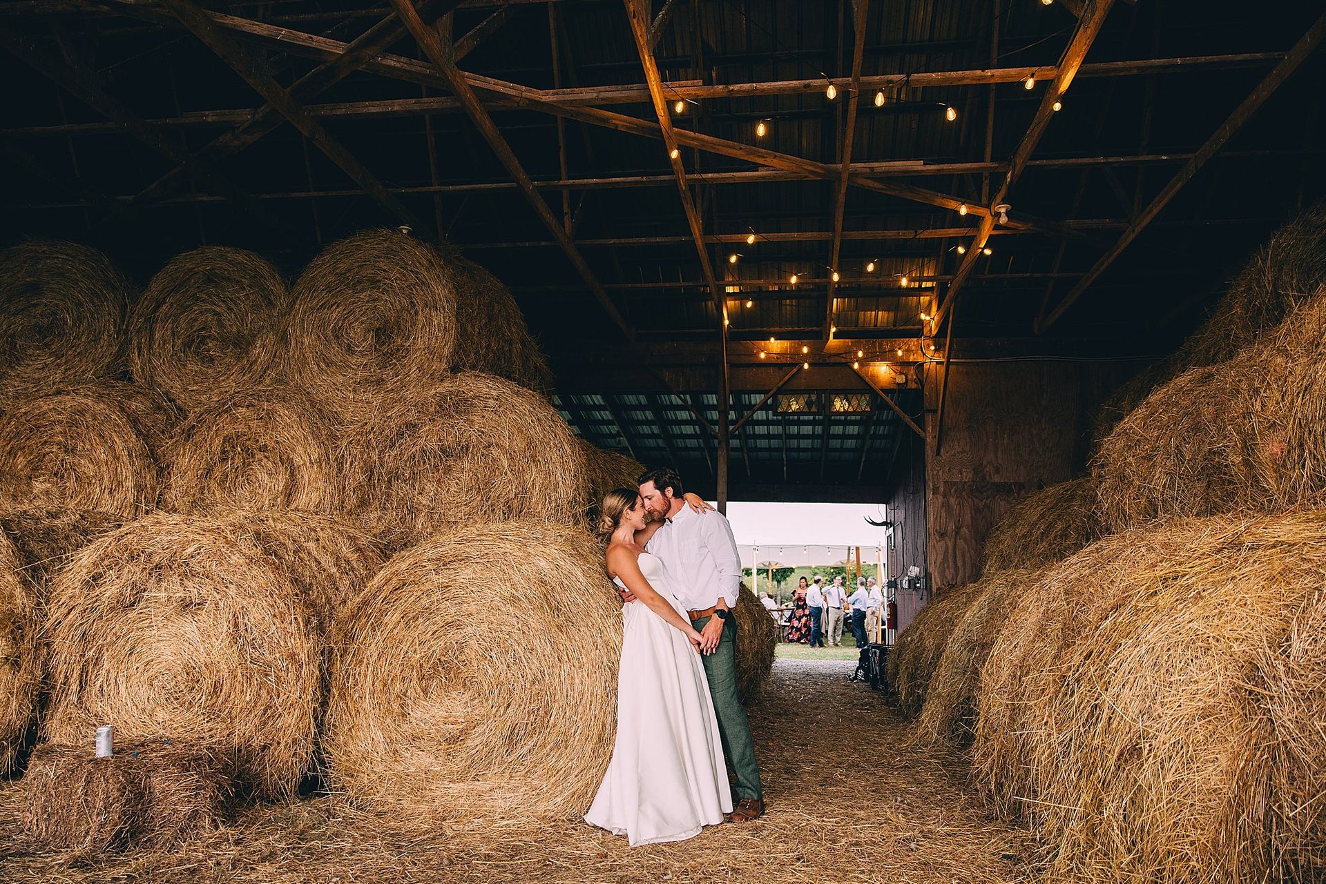 Couple embracing in a barn, surrounded by hay bales, with string lights overhead.