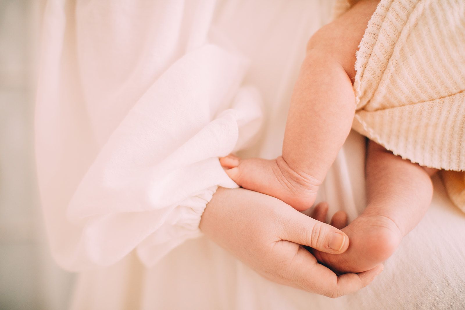 Hands cradle a baby's tiny feet; soft focus, cream-colored clothing and blanket.