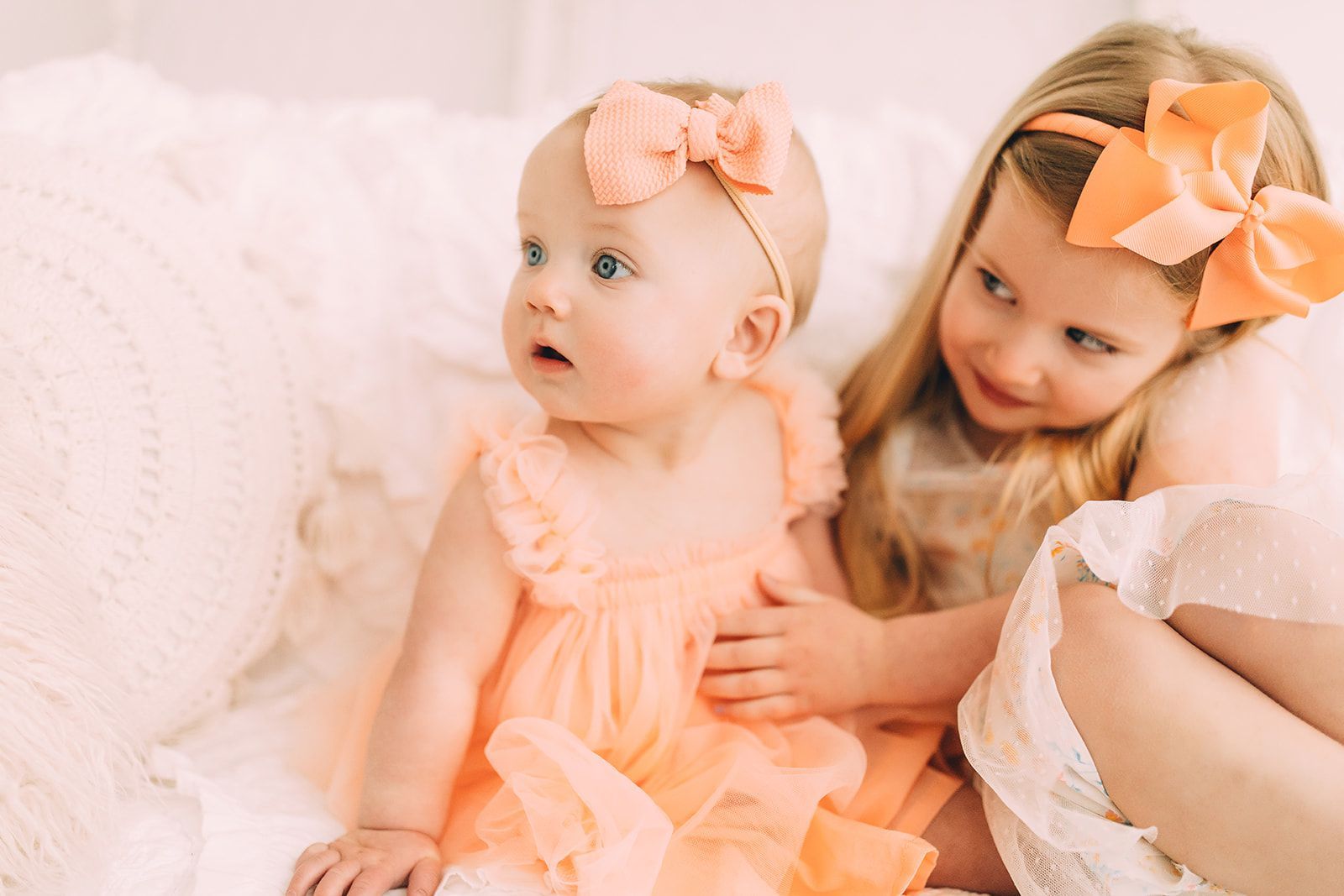 Two girls in peach-colored dresses and bows sitting on a white bed, one looking surprised.