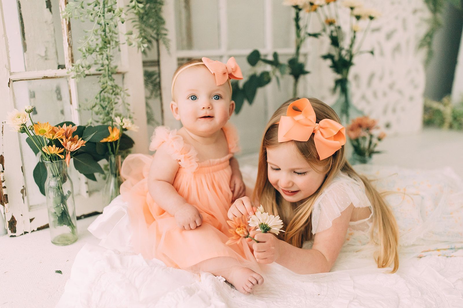 Two girls with coral bows, wearing dresses, smile at the camera, surrounded by flowers.