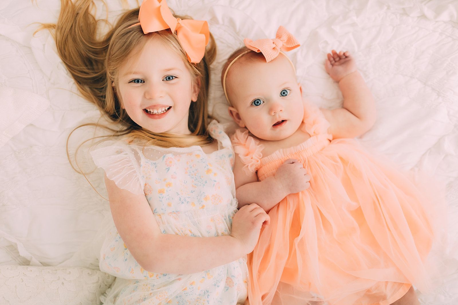 Two young girls, smiling, lying on white bedding; wearing peach bows and dresses.