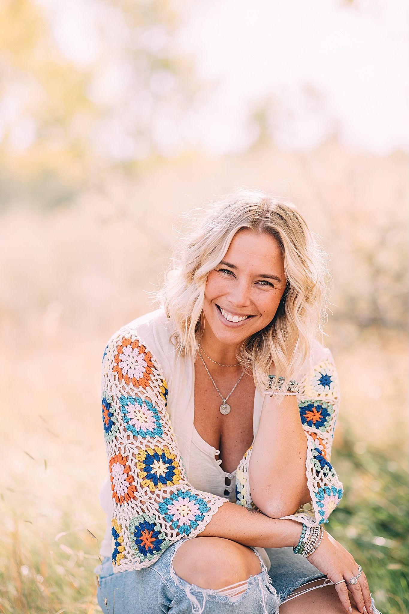 Woman with curly blonde hair smiles outdoors, wearing a crochet top and ripped jeans.