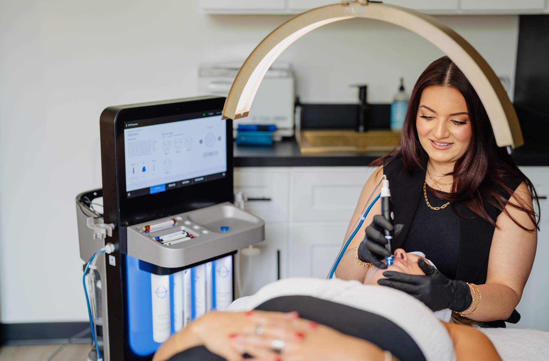 Woman performing facial treatment on a client in a clinic. She uses a device with a screen and light