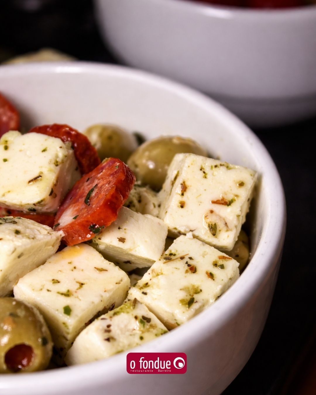 Tigela de cubos de queijo feta marinados com azeitonas e fatias de pimentão vermelho em um prato branco.