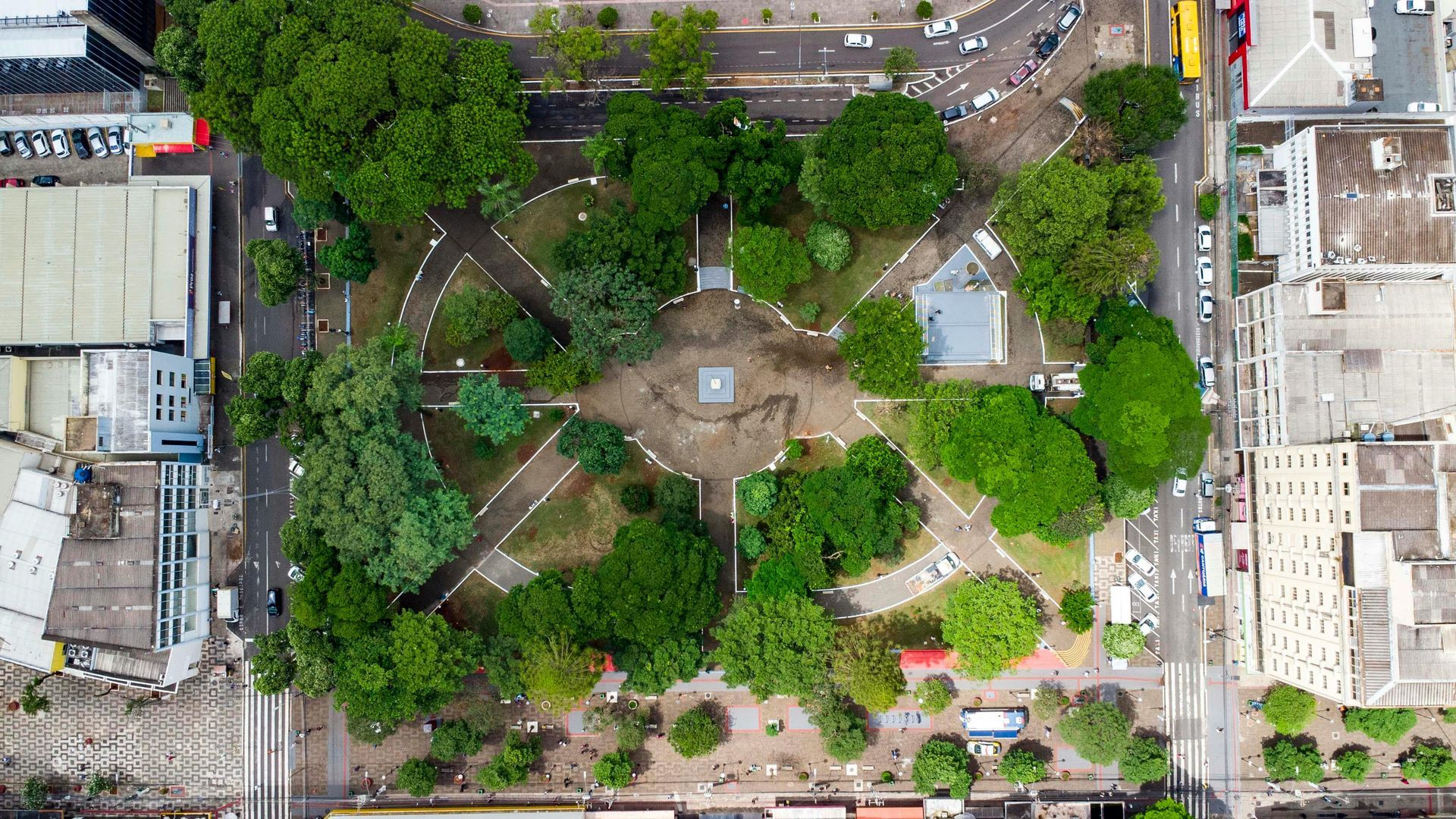 Overhead view of a square park with walkways, trees, and a central monument, surrounded by buildings and streets.