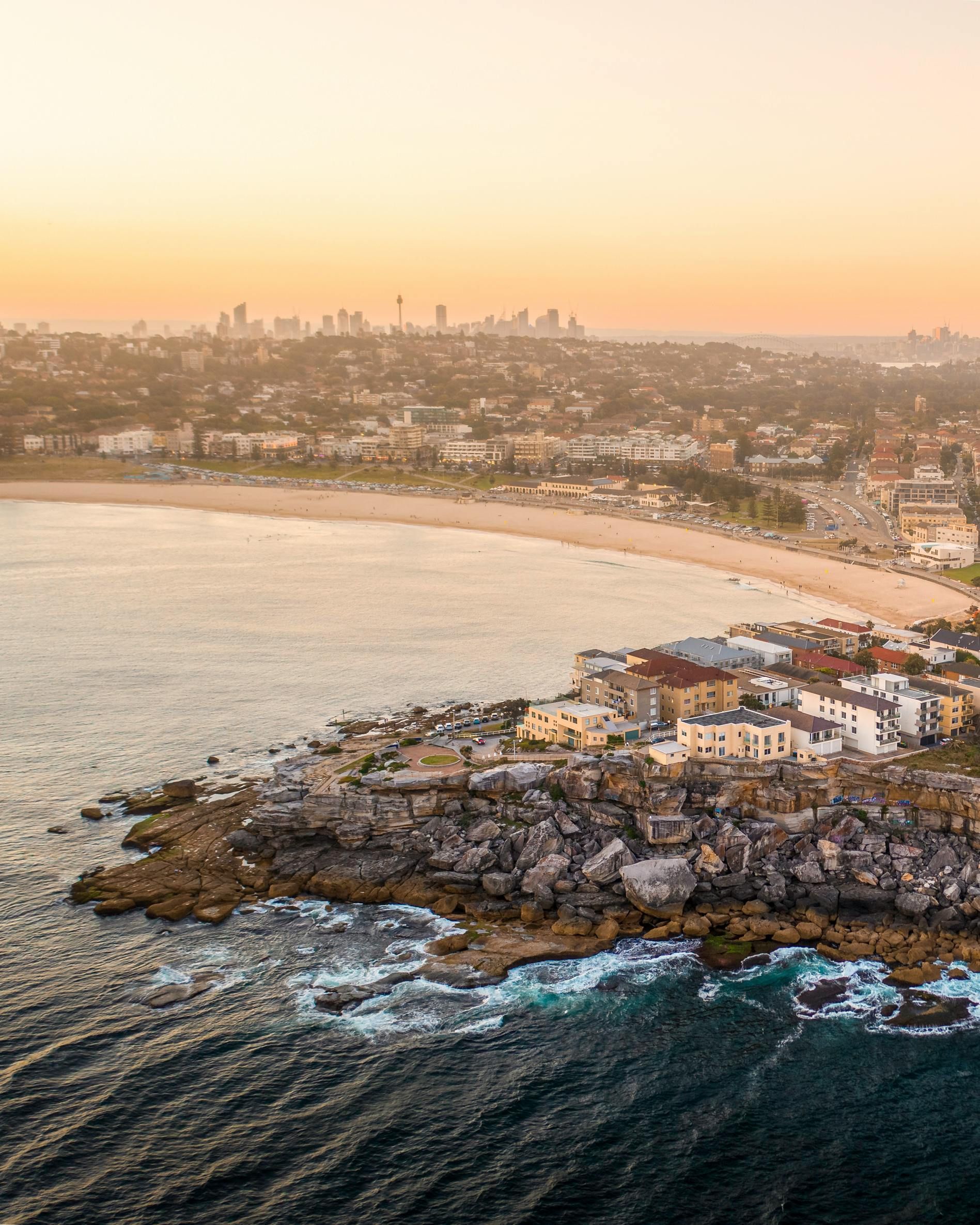 Coastal cityscape at sunset: beach, ocean, buildings on cliffs, distant skyline in orange haze.