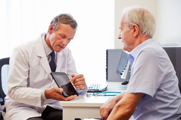 Doctor reviewing medication with a patient at a desk; pills and prescription bottles are visible.