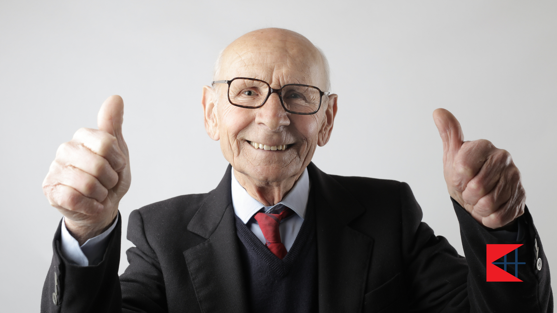 An elderly man in a suit and tie is giving two thumbs up.