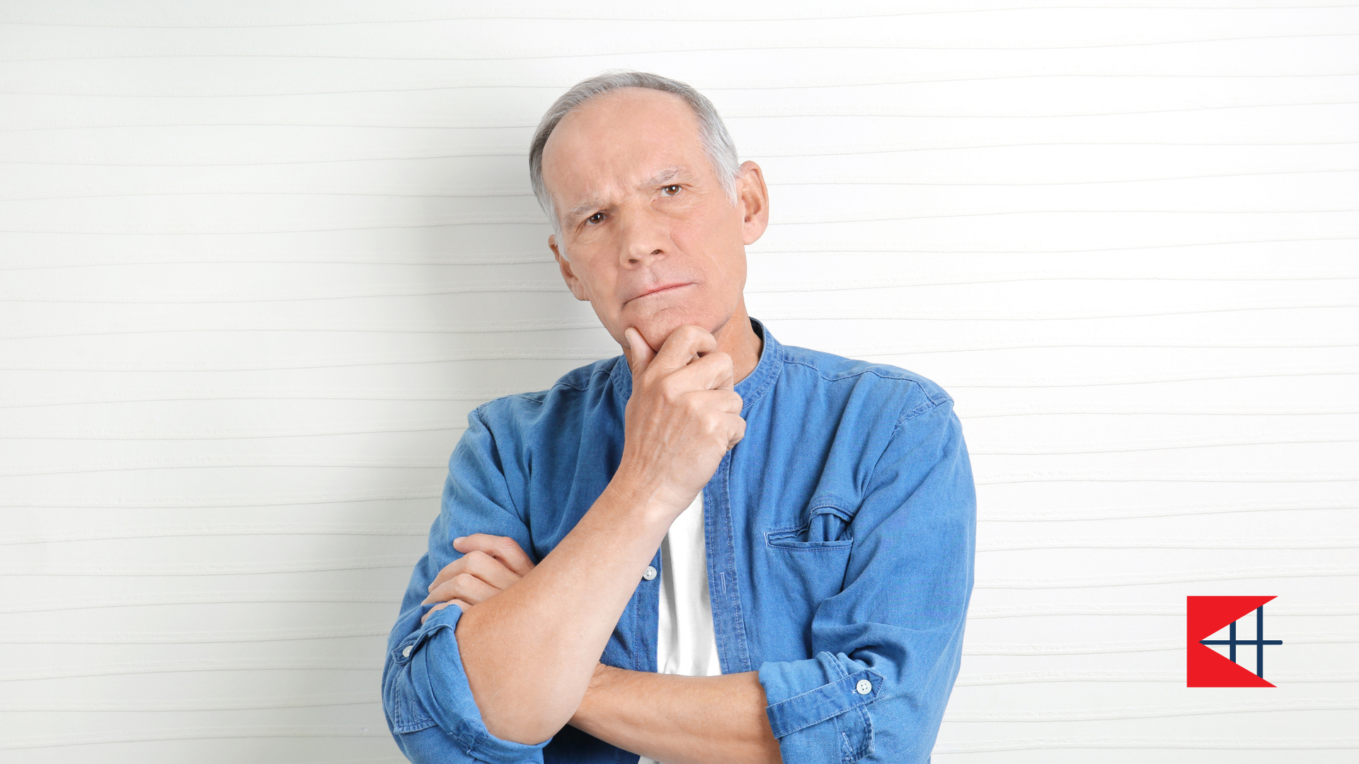 An older man is standing in front of a white wall with his arms crossed and his hand on his chin.