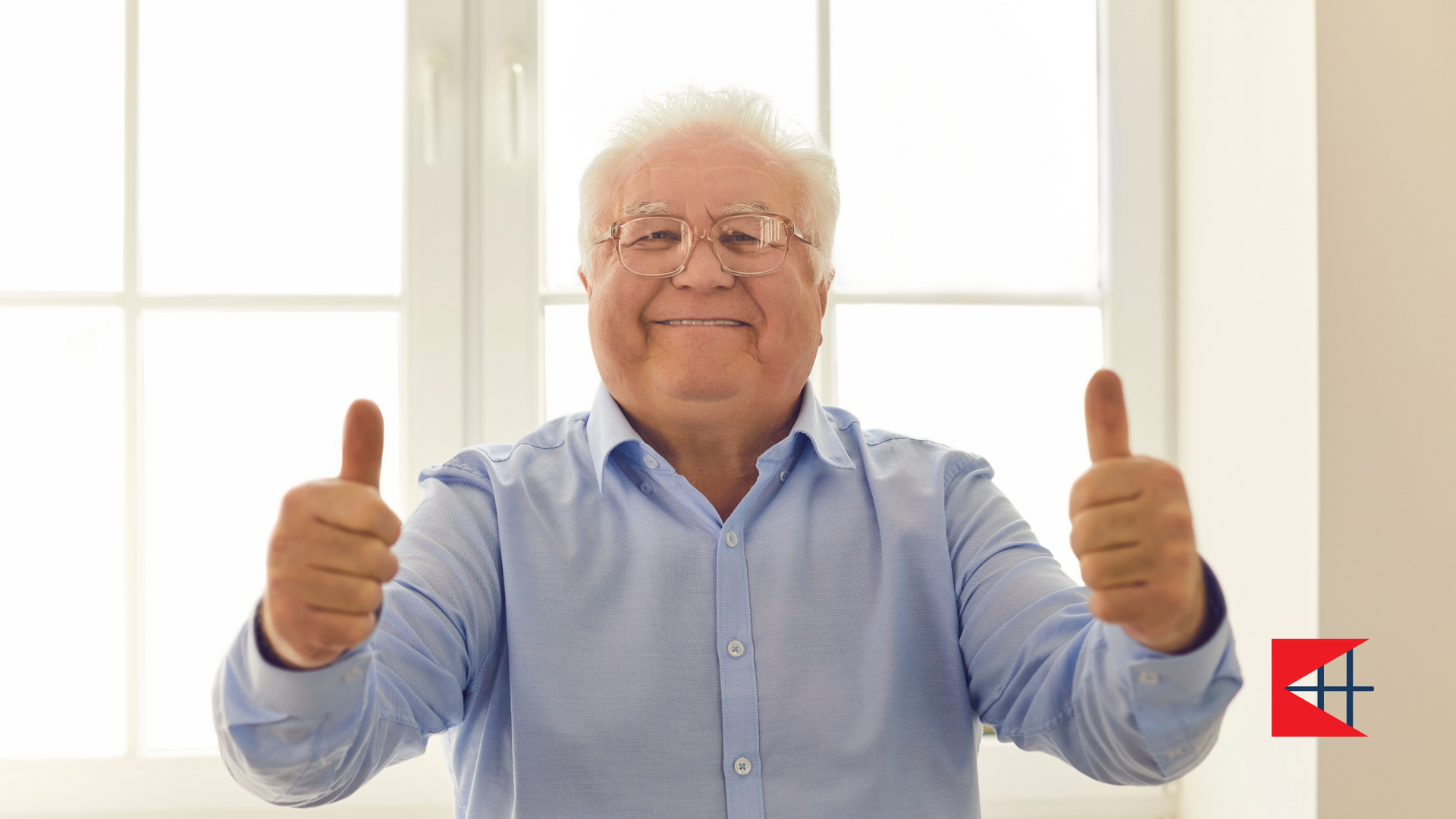 An elderly man is giving two thumbs up in front of a window.