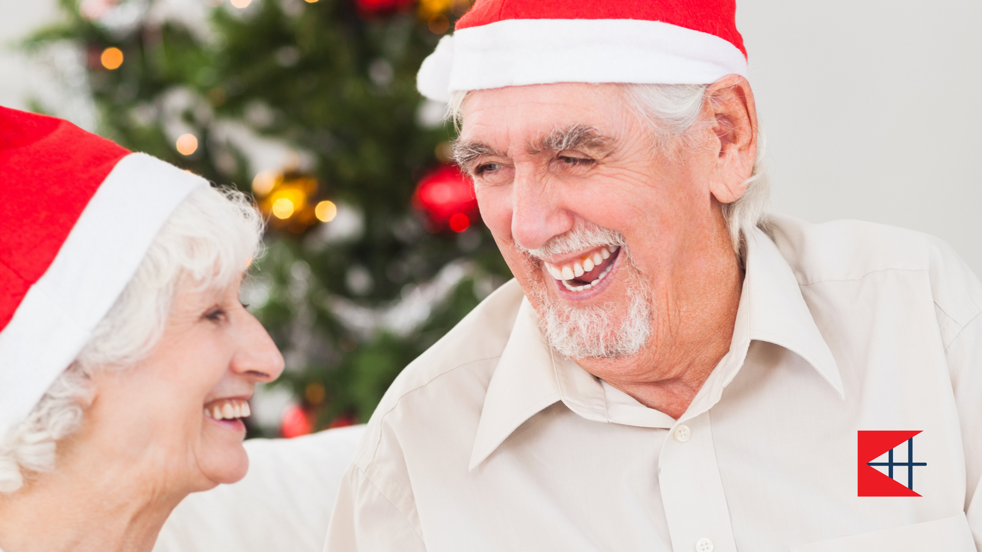 An elderly couple wearing santa hats are smiling in front of a christmas tree.