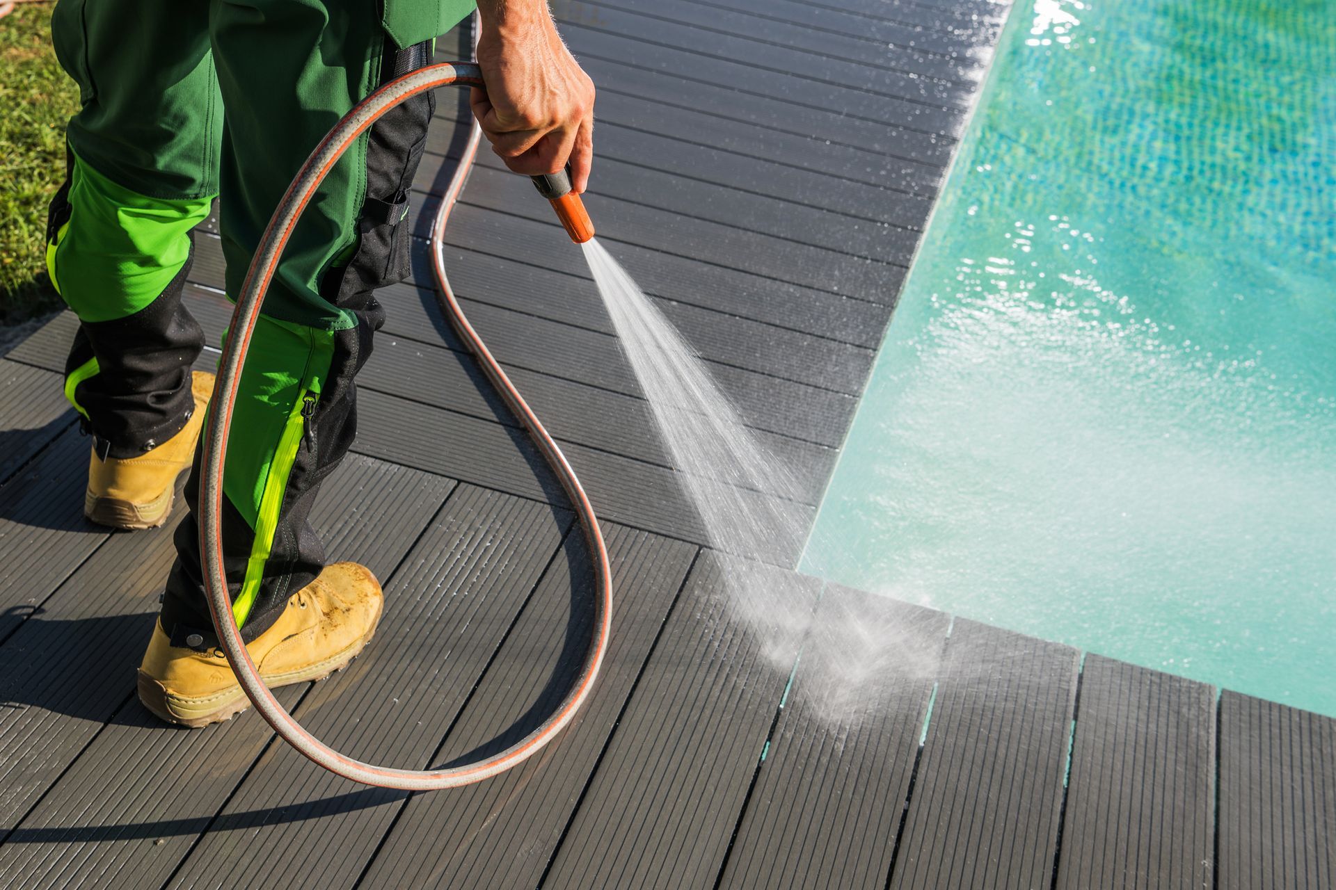 Person sprays water with a hose onto a gray deck next to a swimming pool.