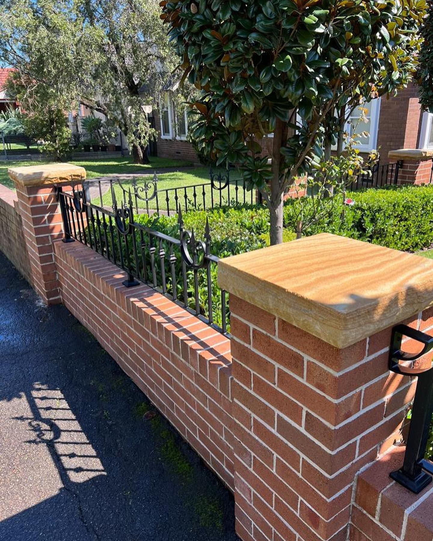 Brick wall with sandstone caps and black iron fence in front of a house, sunny day.