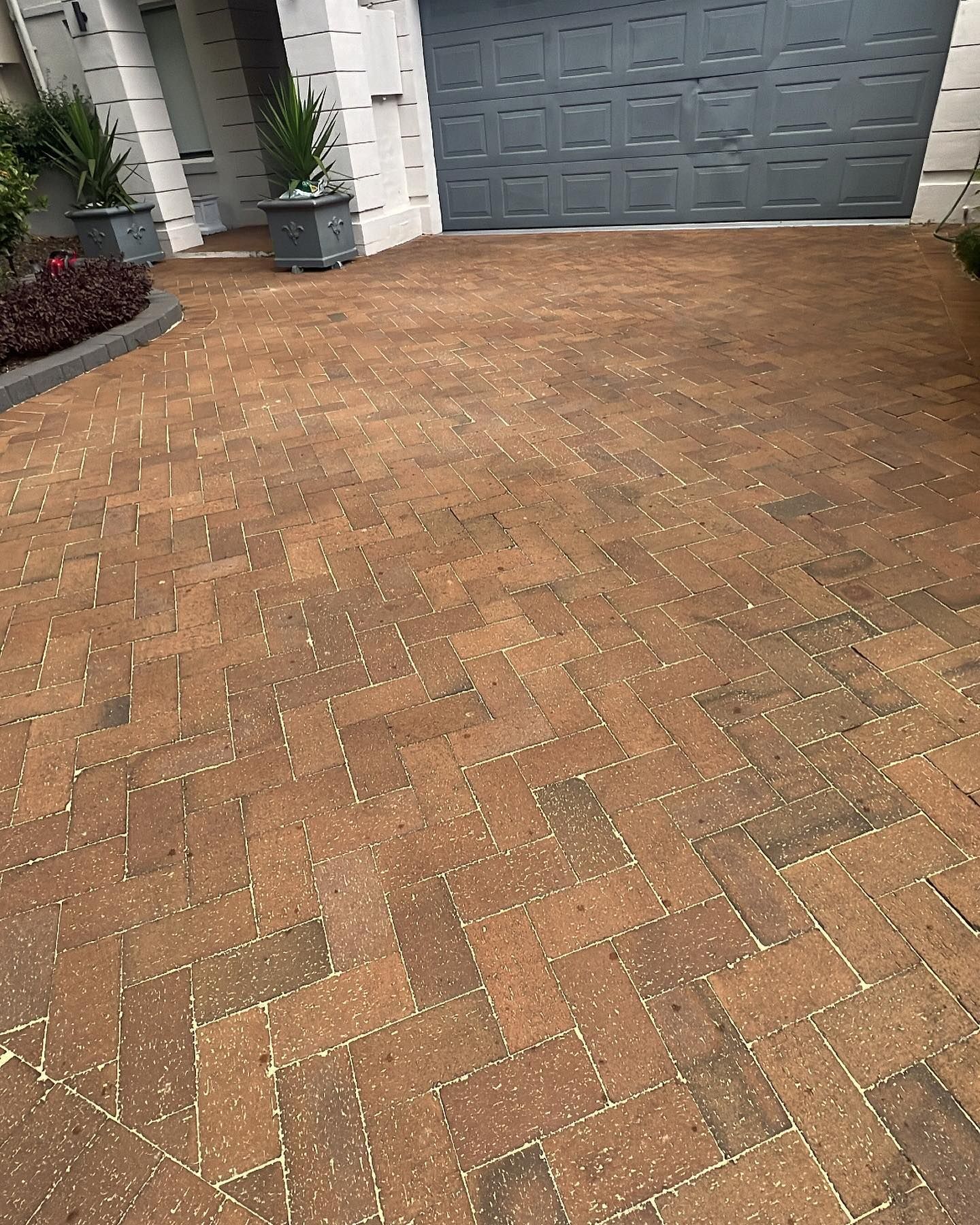 Brick driveway in front of a garage, with potted plants and a small garden.