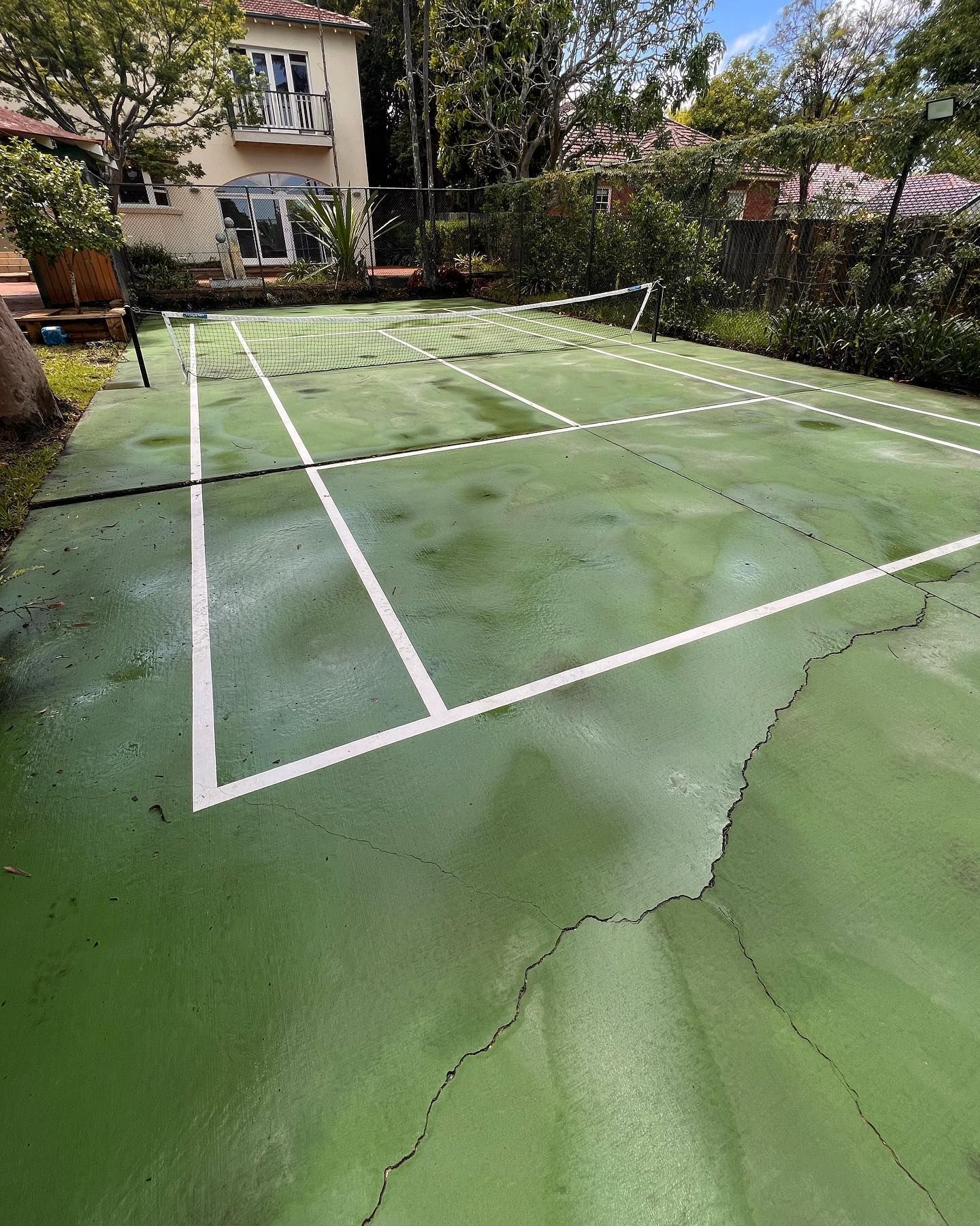 Green tennis court with white lines, net, and a house in the background.