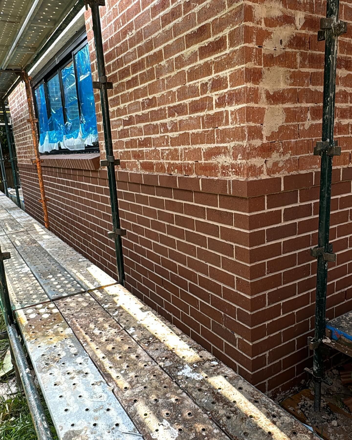 Brick wall under construction, scaffolding and blue-covered window. Red brickwork.