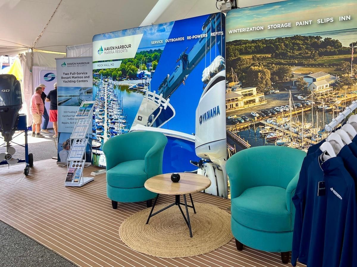 A boat show booth with blue armchairs, a small table and display banners of boats and a harbor