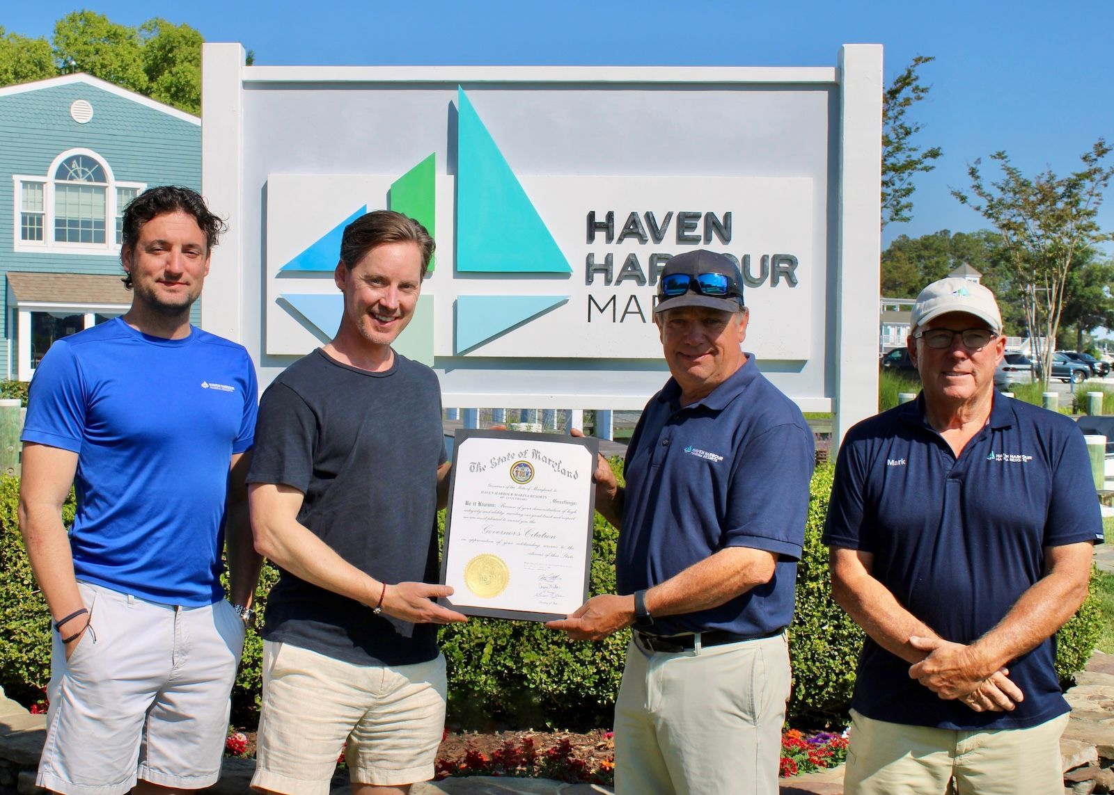 Four men stand holding a certificate in front of a Haven Harbour sign on a sunny day.