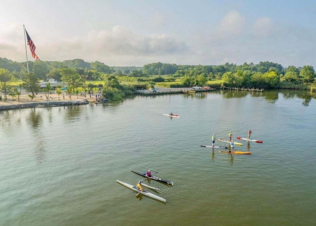 Paddlers and kayakers gather in a harbor with beach in background