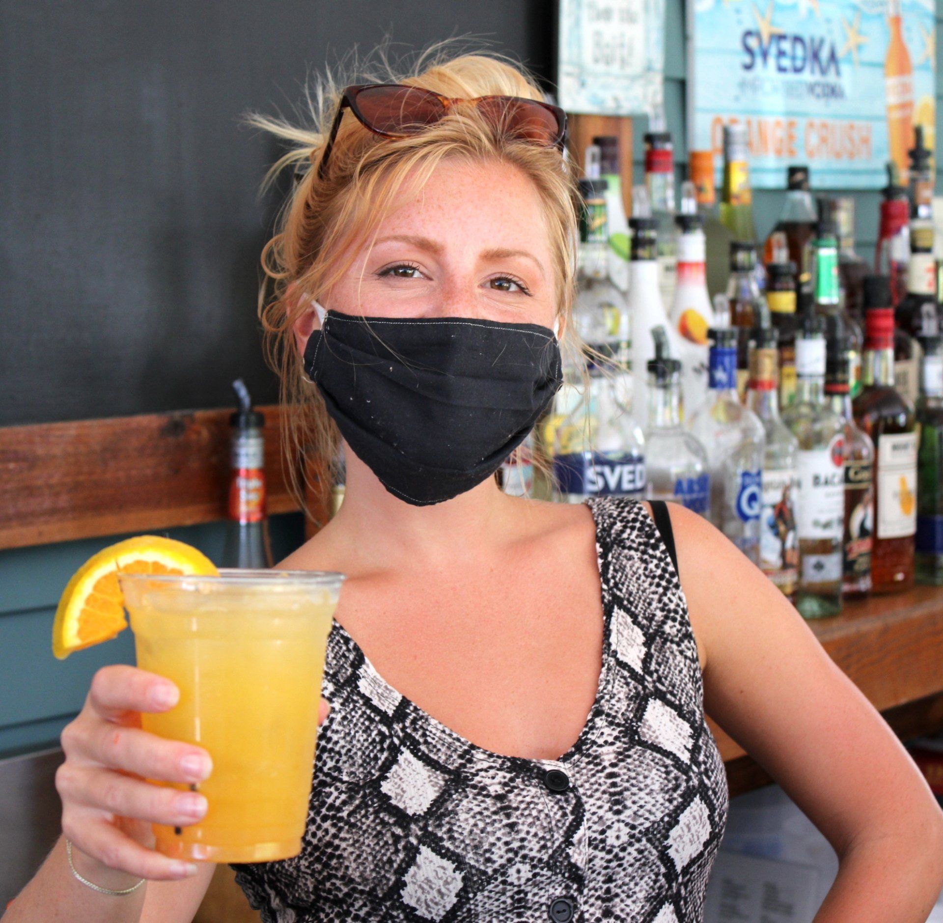 Bartender holds a freshly-made orange cocktail while wearing a mask