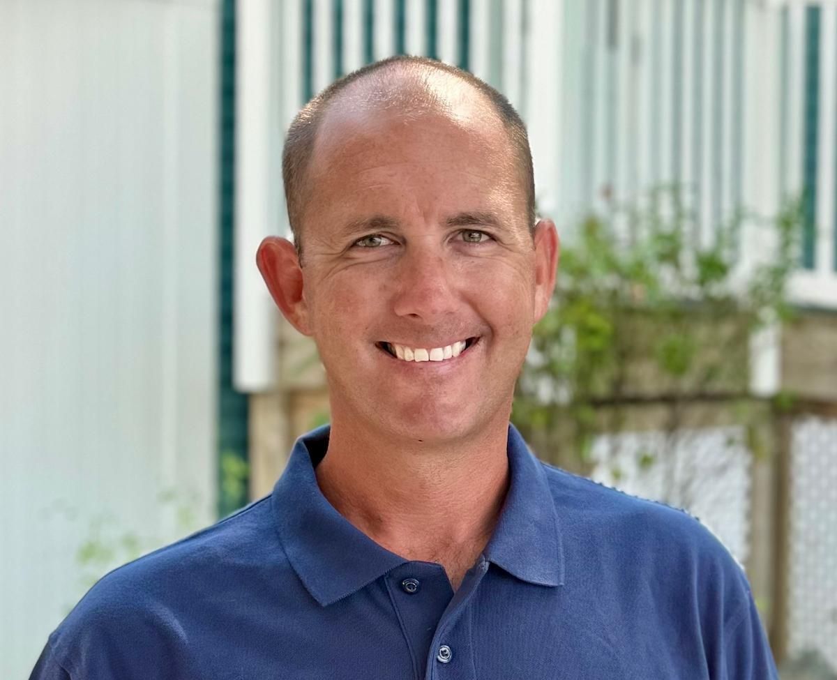 Man posing for photo wearing blue polo shirt in front of greenery in background
