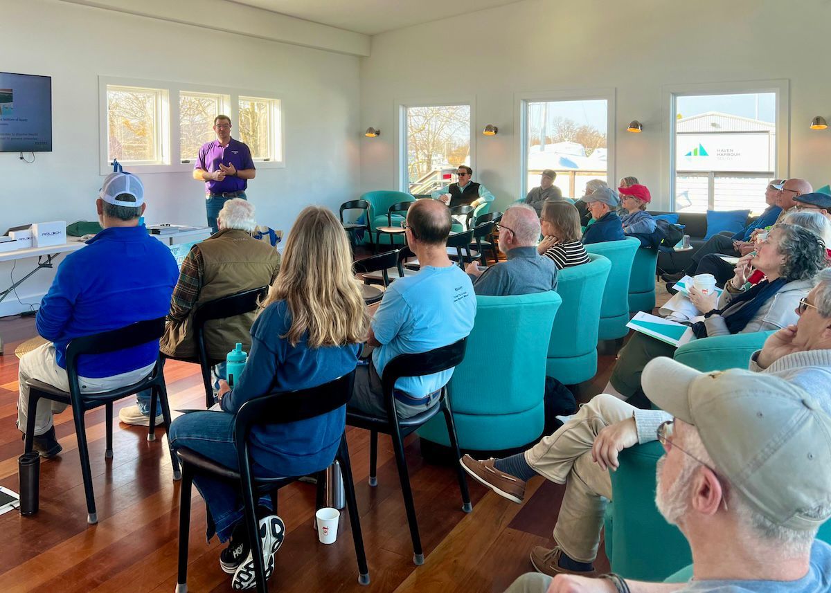 A speaker stands before a seated group in a sunlit room, presenting in front of a monitor.