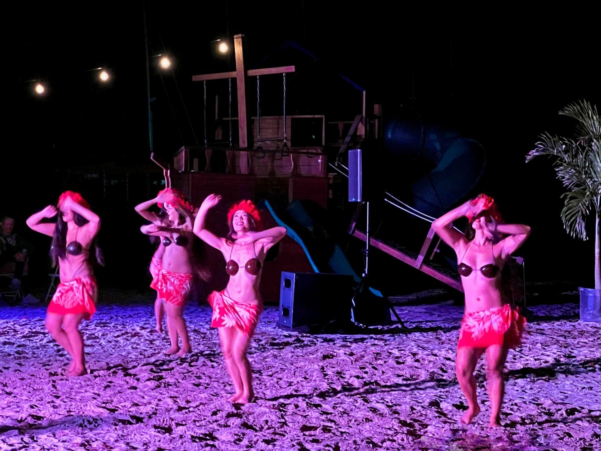 Hula dancers on a beach stage at night; they wear red skirts, shell tops and headdresses
