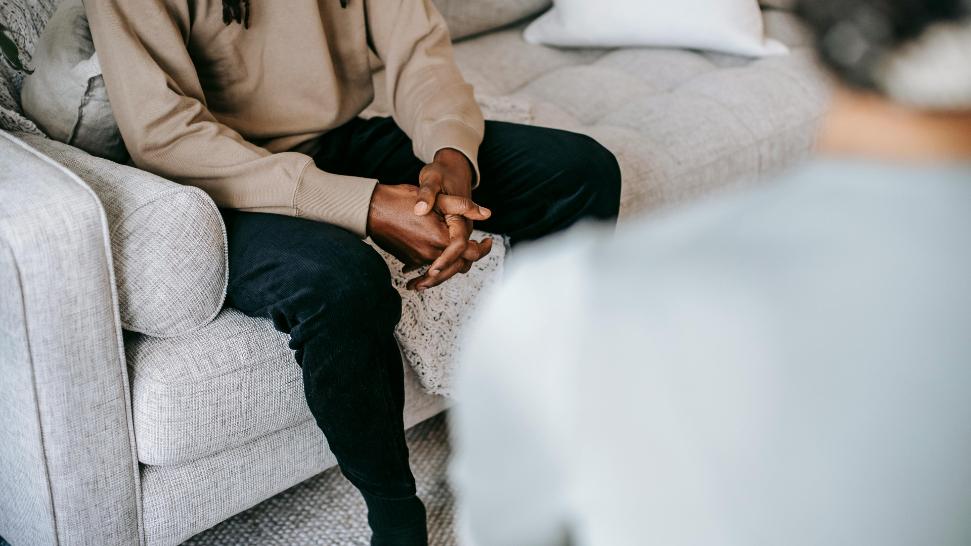 Person with hands clasped sits on a couch, facing another person in a counseling session.